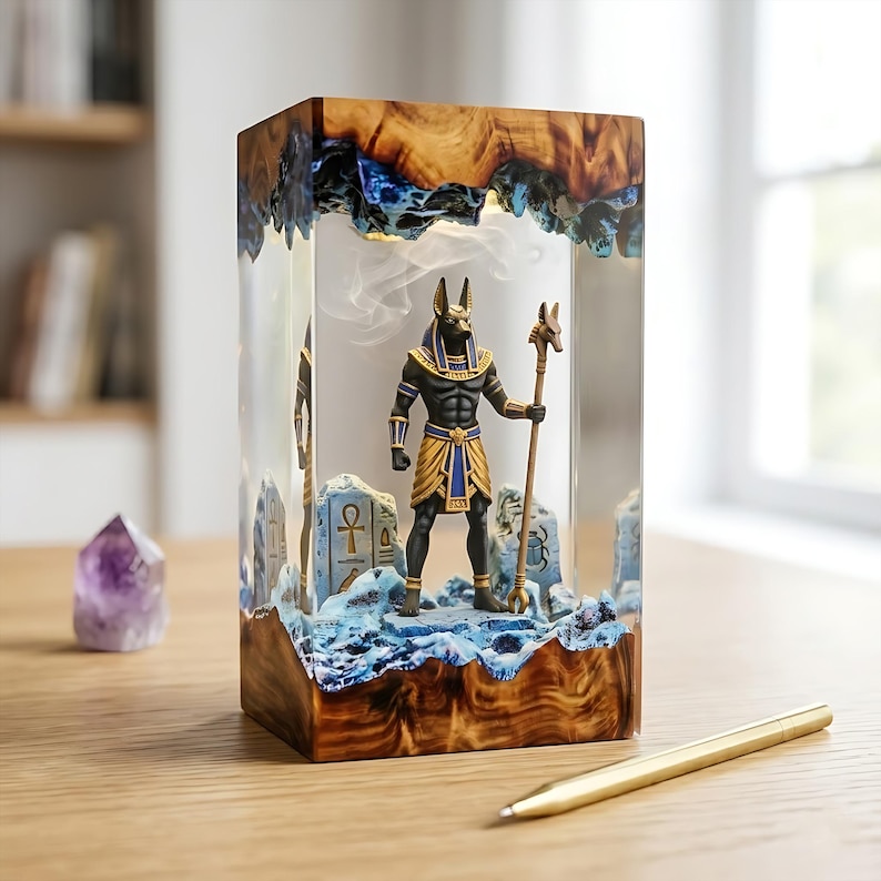May include: A rectangular resin lamp featuring a central Anubis figurine holding a staff, surrounded by blue rocky textures and wood-grain top and bottom sections. The interior displays carved stone tablets with ankh and scarab symbols in a clear block.