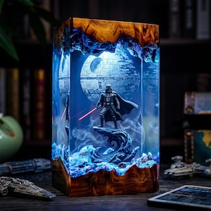 May include: A rectangular resin lamp featuring a wooden top and base with a clear centre containing a miniature Darth Vader figure holding a red lightsaber, standing on a rocky formation in front of a detailed Death Star sphere with blue lighting.