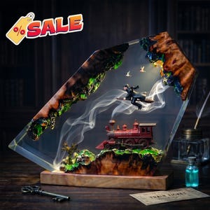 May include: A decorative resin and wood sculpture featuring a miniature red steam locomotive on a rocky base, with a figure flying on a broomstick above it. Wisps of white smoke rise from the train, and two small birds are visible in the air.
