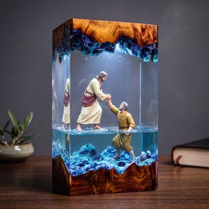 Puede incluir: Una lámpara rectangular de madera natural y resina transparente con una escena de una figura con túnica crema que tiende la mano para sacar a otra figura con túnica beige de un agua de resina azul texturizada. Los bordes son de madera.