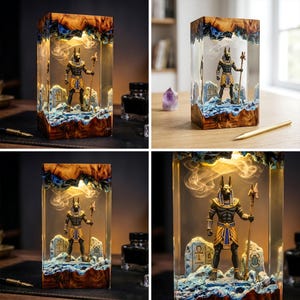 May include: A rectangular resin lamp featuring a miniature Anubis figure standing on a rocky base with blue accents. The figure holds a staff and is flanked by stone carvings of an ankh and a scarab. The top and bottom are encased in wood.