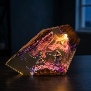 May include: A decorative resin sculpture featuring two samurai figures in combat. The sculpture is illuminated from within, showcasing a fiery, colorful display of orange, pink, and purple hues. The base is a dark wood.
