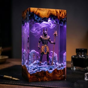 May include: A rectangular resin lamp featuring a central figurine of the Egyptian deity Anubis holding a staff. The lamp is encased in clear resin with a wood-grain top and base, illuminated by a purple light with swirling smoke-like effects.