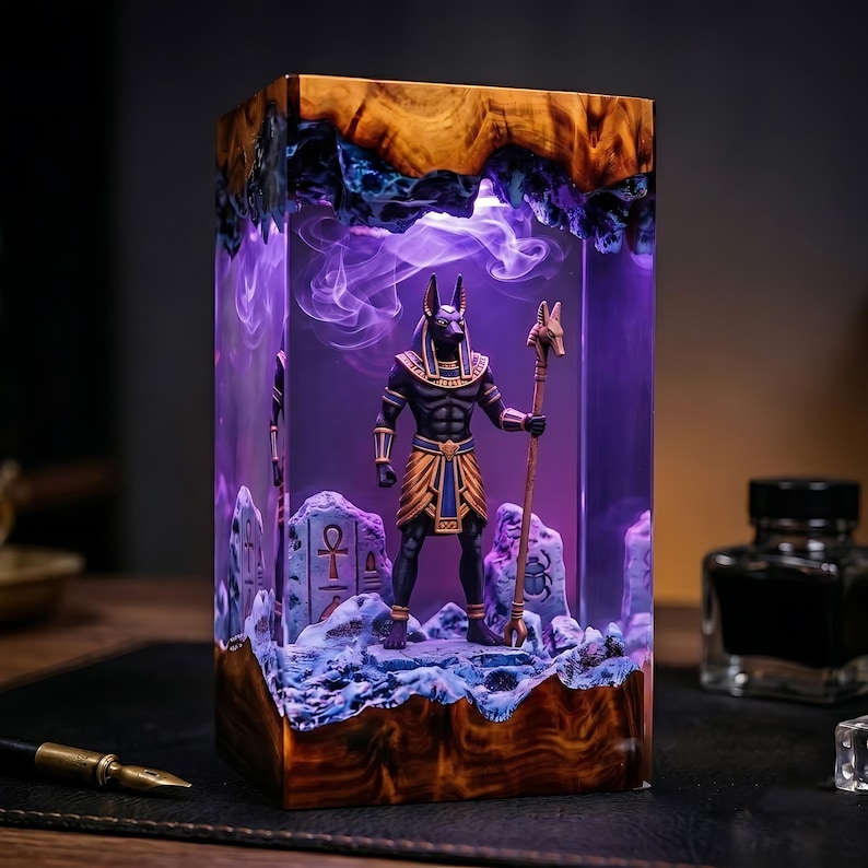 May include: A rectangular resin lamp featuring a central figurine of the Egyptian deity Anubis holding a staff. The lamp is encased in clear resin with a wood-grain top and base, illuminated by a purple light with swirling smoke-like effects.