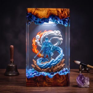 Op de afbeelding: Een rechthoekige harslamp met een doorschijnend blauw krijgersfiguur dat een staf vasthoudt, omringd door gebeeldhouwde oranje vlammen en witte wervelende wolken. De voet en de bovenkant zijn gemaakt van gepolijst hout met ledverlichting.