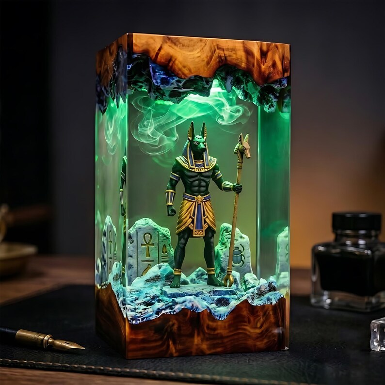 May include: A rectangular resin lamp featuring a wooden base and top, enclosing a green-lit diorama of the Egyptian deity Anubis holding a staff. The scene includes miniature stone-like carvings with hieroglyphs and a scarab beetle symbol.