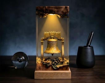 May include: A Liberty Bell sculpture encased in clear resin, illuminated by a warm light. The bell is gold-colored, with a crack visible. The base is wood, and the top of the resin has a dark, textured appearance. A pen and a glass sphere are on the wooden surface.
