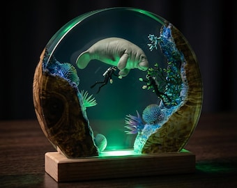 May include: A handcrafted resin sculpture featuring a manatee and a scuba diver within a clear, rounded enclosure. The sculpture includes simulated coral and plant life, with a wooden base and a green LED light.