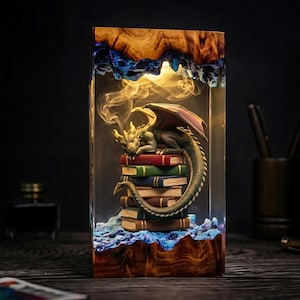 May include: A rectangular resin lamp featuring a carved wood top and base with a blue textured interior. Inside, a green dragon with wings rests atop a stack of six colorful books. The lamp emits a warm light, creating a smoke-like effect.