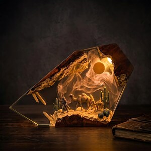 May include: A decorative lamp featuring a desert scene with camels, a figure, and cacti. The lamp has a wooden and resin frame with a warm light emanating from within, creating a sunset effect. The base is a gold metal frame.