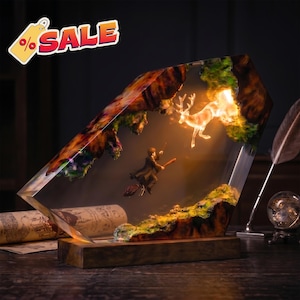 May include: A resin lamp featuring a miniature figure riding a broomstick and a glowing stag patronus. The scene is encased in a clear, angular block with dark, textured edges and sits on a rectangular wooden base. A sale tag is visible.