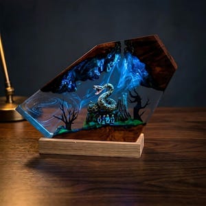 May include: A decorative resin sculpture featuring a detailed snake, miniature figures, and stylized trees. The sculpture is encased in clear resin with blue and white swirls, set on a wooden base. The wood has a dark brown grain.