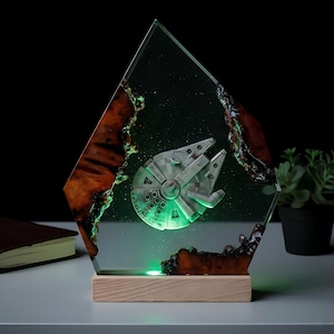 Peut inclure: Une œuvre d'art en résine sur le thème de Star Wars représentant le vaisseau spatial Millennium Falcon. L'œuvre est enfermée dans un bloc de résine clair et géométrique avec une base en bois. La résine a une lueur verte et les bords sont bordés d'un matériau sombre et texturé.