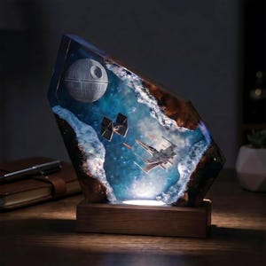 May include: A Star Wars resin lamp depicting the Death Star, X-wing fighters, and TIE fighters in a space scene. The lamp sits on a wooden base and provides a soft, ambient light. The scene is encased in a clear resin.