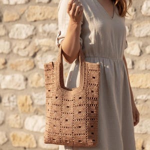 Op de afbeelding: Een bruin gehaakte tote bag met een rechthoekige vorm en een open weefselontwerp. De tas heeft een stevige handgreep en wordt vastgehouden door een persoon die een lichtgekleurde jurk draagt. De achtergrond is een stenen muur.