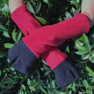 May include: Red and black fleece gloves with a split-toe design. The gloves are red with black tips and are shown against a background of green foliage. The gloves appear to be warm and comfortable.