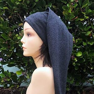 May include: A dark gray knit hat with a long, pointed tail and a tassel. The hat is displayed on a mannequin head against a backdrop of green foliage. The hat's design is simple and elegant.