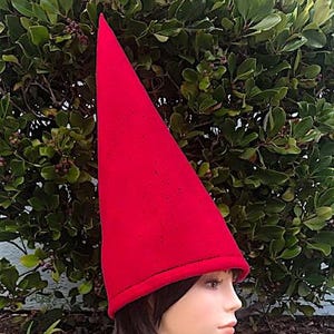 May include: A bright red, cone-shaped hat with a slightly textured surface. The hat has a dark band around the base. The hat is on a mannequin head with short, dark hair. The background is a green bush.