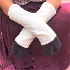 May include: White and black plush leg warmers. The leg warmers are white with black hooves. The person is wearing a purple patterned top and sitting on a purple surface.