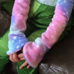 May include: A pair of fleece arm warmers in a galaxy print, featuring shades of pink, blue, and purple with white star accents. The arm warmers have a heart-shaped design at the hand. The person is wearing a green outfit.