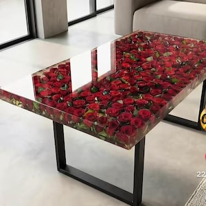 May include: A rectangular coffee table with a clear resin top filled with numerous red roses. The table has a black metal frame and is set in a modern living room setting.