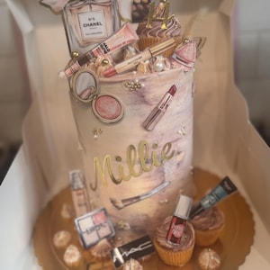 May include: A birthday cake decorated with makeup-themed toppers, including a perfume bottle, lipstick, and eyeshadow. The cake has a marble effect with the name "Millie" in gold lettering. Cupcakes with frosting and makeup decorations are also present.