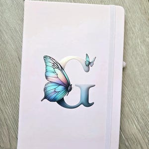 Può includere: Un diario rosa chiaro con una farfalla e la lettera "G". La farfalla ha ali blu, viola e rosa. Una piccola farfalla è accanto alla lettera "G". Il diario ha una chiusura con elastico bianco.