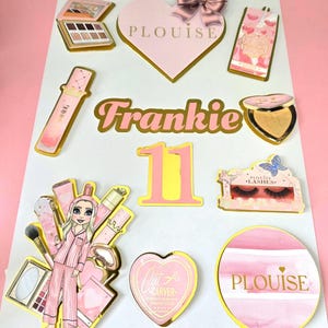 Op de afbeelding: Een verzameling roze en gouden make-up-thema decoraties, waaronder een hart met een strik, een lippenstift en een palet. De naam "Frankie" en het nummer "11" zijn ook te zien. De decoraties zijn gerangschikt op een wit oppervlak.