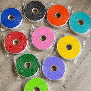 Op de afbeelding: Tien ronde rollen zelfklevende schuimrubberen tochtstrip, elk afzonderlijk verpakt in doorzichtig plastic. De rollen zijn verkrijgbaar in verschillende effen kleuren, waaronder zwart, blauw, oranje, turquoise, rood, roze, geel, groen, paars en limoengroen.