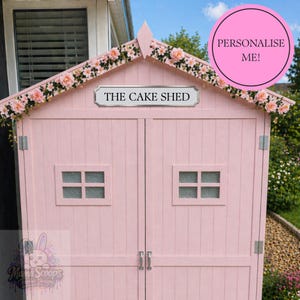 Może przedstawiać: Różowa drewniana szopa z dwuspadowym dachem, ozdobiona różowymi kwiatami. Znak głosi "THE CAKE SHED". Szopa ma dwa małe okna i podwójne drzwi. Widoczny jest różowy okrąg z napisem "PERSONALIZE ME!".