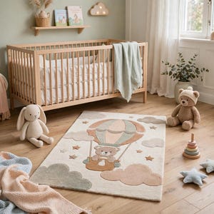 Op de afbeelding: Een rechthoekig vloerkleed met een centrale illustratie van een teddybeer in een heteluchtballon, omringd door wolken en sterren in gedempte tinten beige, saliegroen en zachtbruin op een crèmekleurige achtergrond.