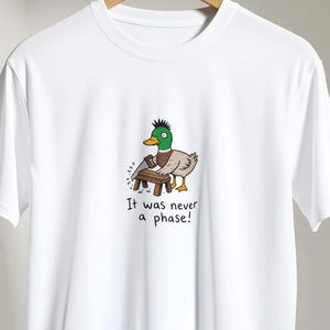 May include: A white short-sleeved t-shirt featuring a graphic of a mallard duck with a green mohawk hairstyle, wearing a brown collar, and standing at a small wooden workbench. The text below the duck reads, It was never a phase!
