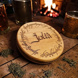 May include: A round, light-colored wooden box lid featuring a wood-burned inscription of the name Julia in the center, surrounded by a circular border of engraved elvish-style script, resting on a wooden surface with scattered dried herbs.