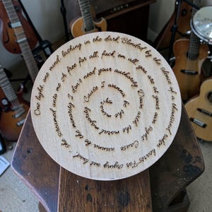 Peut inclure: Un objet décoratif rond en bois avec du texte gravé en spirale. Le texte comprend des phrases comme "lashing in the dark" et "I wanna be the minority." Des guitares sont visibles en arrière-plan.