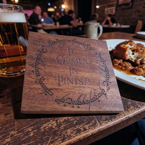 Puede incluir: Un posavasos cuadrado de madera marrón con el texto "It Comes in Pints?!" en un diseño circular. El posavasos está sobre una mesa de madera. Un vaso de cerveza y un plato de comida se ven en el fondo.