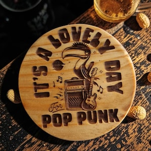 Op de afbeelding: Een ronde houten onderzetter met de tekst "IT'S A LOVELY DAY For POP PUNK" en een toekan illustratie. De onderzetter heeft ook een versterker en gitaar ontwerp, met muzieknoten. Pinda's omringen de onderzetter.