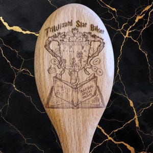 May include: A wooden spoon with a laser-etched design. The design features the text "Triwizard Star Baker" above a trophy and an open book with the words "Baking Spells Treats". The spoon is light brown and has a long handle.