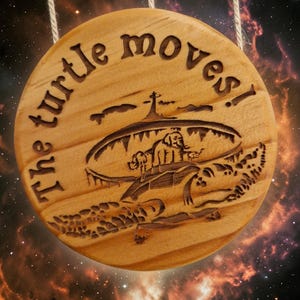 May include: Round wooden sign with the text "The turtle moves!" carved into the surface. The sign features a detailed engraving of a turtle carrying a structure on its back. The sign is suspended by a natural fiber cord.