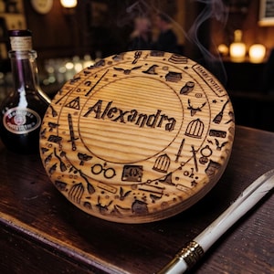 Può includere: Un oggetto decorativo rotondo in legno con il nome "Alexandra" inciso al centro, circondato da simboli e testo incisi di Harry Potter. Sono visibili anche una bottiglia di liquido scuro e una penna.