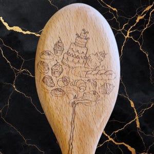 May include: A wooden spoon with a detailed pyrography design featuring various baked goods, including a tiered cake, cupcakes, and pastries. The spoon is made of light-coloured wood and is set against a black and gold marble background.
