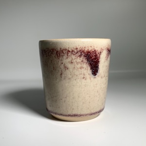 May include: A handmade ceramic cup with a cream base and a speckled, textured surface. The rim and upper part of the cup have a deep burgundy glaze that drips down the sides. The cup has a simple, cylindrical shape.
