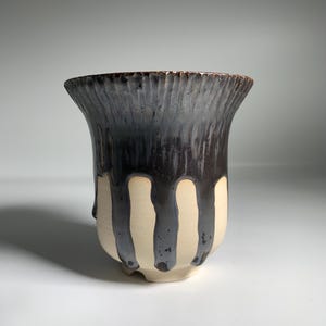Può includere: Una tazza in ceramica fatta a mano con un design unico. La metà superiore è marrone scuro con scanalature verticali, mentre la metà inferiore è color crema con gocciolature marroni scure. La tazza ha un bordo svasato e una base arrotondata.