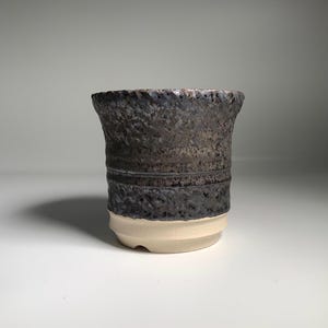 Op de afbeelding: Een handgemaakte keramische beker met een getextureerd, donkergrijs bovenstuk en een donkerblauwe band. Het onderste deel is crèmewit van kleur. De beker heeft een licht uitlopende rand en een stevige basis.