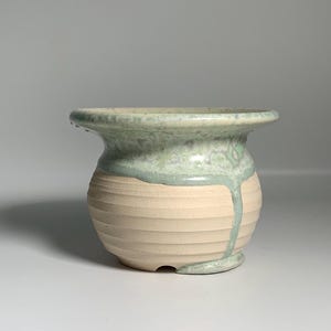 Puede incluir: Pequeño jarrón de cerámica hecho a mano, con cuerpo beige claro y esmalte verde claro. El jarrón tiene forma redondeada con estrías horizontales y un borde ensanchado. El esmalte verde gotea por el lateral.