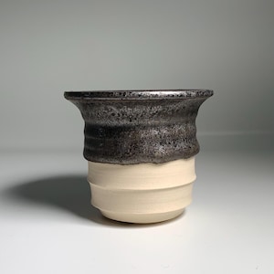May include: A handmade ceramic cup with a dark, textured rim and a cream-colored, ridged body. The cup has a unique, organic shape and a contrasting color scheme, showcasing the craftsmanship of the pottery.