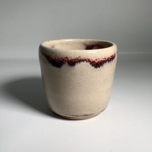 May include: A small, handmade ceramic cup with a rounded shape. The cup is a creamy off-white colour with a dark burgundy rim. The interior and exterior have a speckled texture, giving it a rustic appearance. The cup is suitable for beverages.
