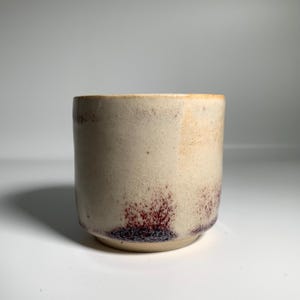 May include: A handmade ceramic cup with a cylindrical shape. The cup has a cream-coloured exterior with reddish-brown speckles near the base. The rim has a light brown colour. The cup is likely used for drinking beverages.