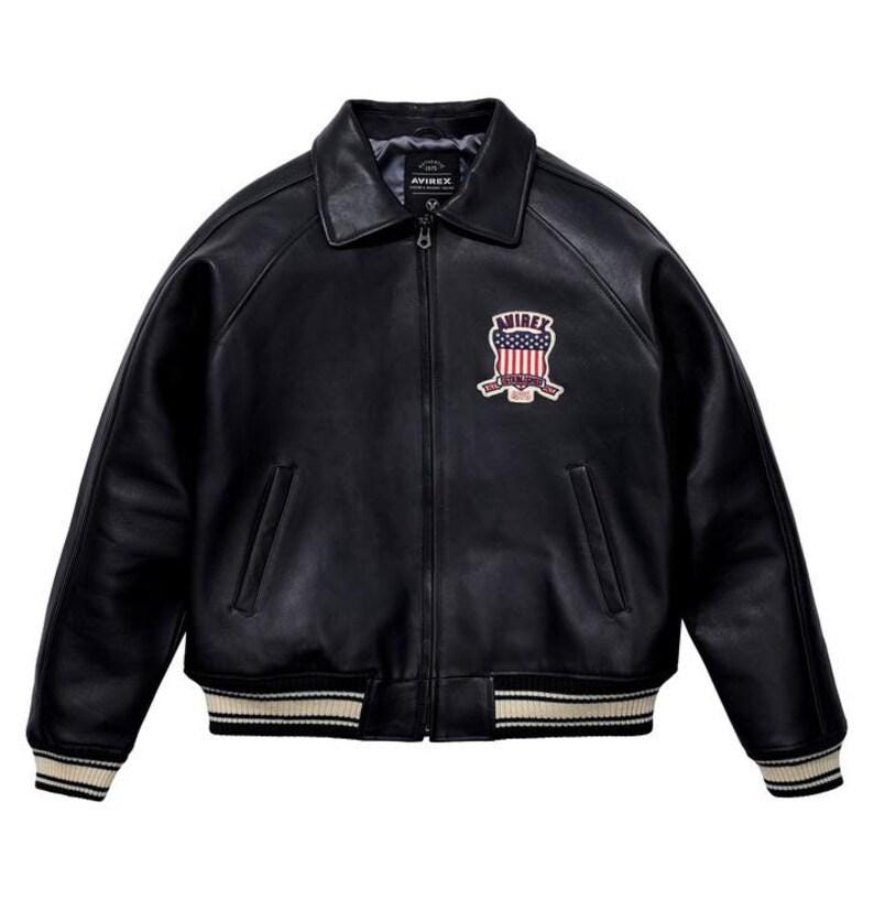 K&ouml;nnte beinhalten: Schwarze Leder-Bomberjacke mit Rei&szlig;verschluss, Kragen und gerippten B&uuml;ndchen und Saum. Verf&uuml;gt &uuml;ber einen Aufn&auml;her auf der Brust mit dem Wort "AVIREX" und einem Design in Rot, Wei&szlig; und Blau. Die Jacke hat zwei Vordertaschen.