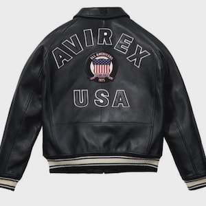 K&ouml;nnte beinhalten: Schwarze Lederjacke mit wei&szlig;em Schriftzug "AVIREX" und "USA". Die Jacke hat einen runden Aufn&auml;her mit der amerikanischen Flagge und der Aufschrift "ALL AMERICANS ESTABLISHED 1975". Sie hat einen gerippten Saum und B&uuml;ndchen.