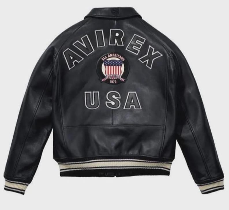K&ouml;nnte beinhalten: Schwarze Lederjacke mit wei&szlig;em Schriftzug "AVIREX" und "USA". Die Jacke hat einen runden Aufn&auml;her mit der amerikanischen Flagge und der Aufschrift "ALL AMERICANS ESTABLISHED 1975". Sie hat einen gerippten Saum und B&uuml;ndchen.